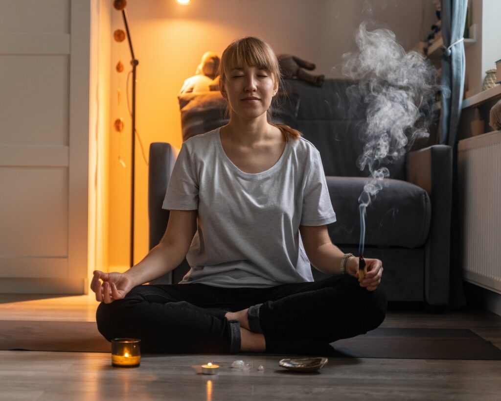 Smiling woman holding aromatic smolding stick while meditating in lotus pose at home