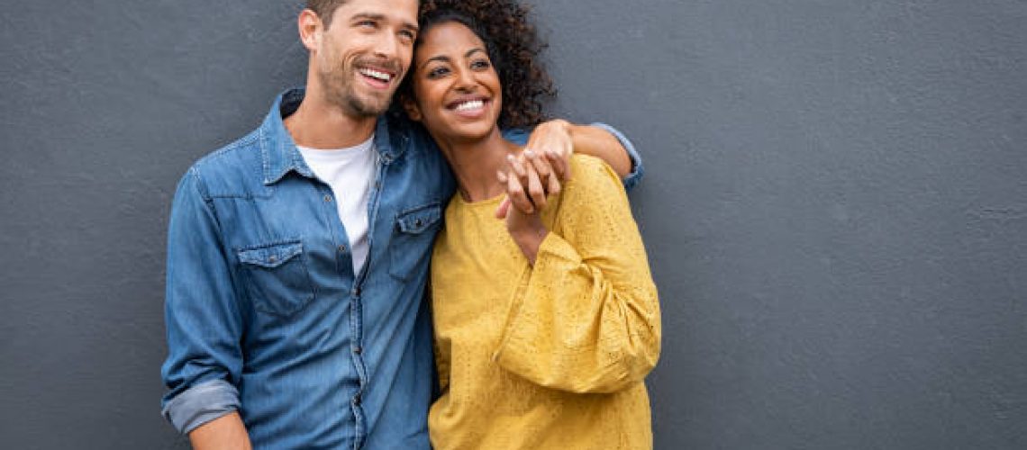 Young multiethnic couple in love isolated on grey background looking up and thinking about their future together. Smiling man and african woman in casual hugging and looking away while planning the future. Cheerful couple holding hands while leaning on wall.
